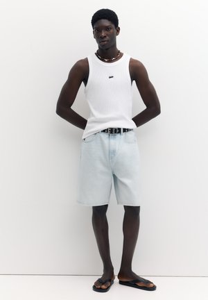 Jeune homme portant un débardeur blanc, un short en jean clair avec une ceinture noire, des sandales et un collier de perles, debout devant un fond blanc.