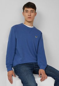 Giovane uomo seduto su un blocco bianco, indossando una felpa blu con una piccola toppa e jeans blu scuro, sfondo grigio neutro.