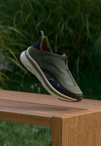 Πράσινα και μαύρα παπούτσια τύπου slip-on με υφή στην επάνω επιφάνεια, κλείσιμο με φερμουάρ και λευκή σόλα, τοποθετημένα σε ξύλινη επιφάνεια με φόντο γρασίδι.