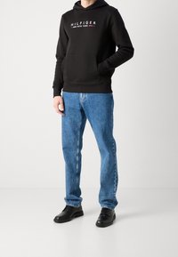 Svart hoodie med vit text "HILFIGER NEW YORK", kombinerad med blå denimjeans och svarta skor. Avslappnad passform, slät textur och minimalistisk design.