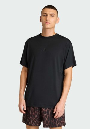 Mand iført sort Adidas t-shirt og mørke mønstrede shorts, står mod en ensfarvet lys baggrund, vendt mod fronten med neutralt ansigtsudtryk.