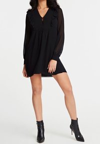Robe noire avec manches longues en voile transparent, col en V et détails à volants. Dotée d'une fermeture à boutons à l'avant. Associée à des bottines noires pointues.
