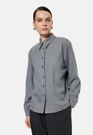 Mujer con el pelo peinado hacia atrás, lleva pendientes de aro plateados y una camisa gris de manga larga abotonada con pantalones gris oscuro sobre un fondo liso.