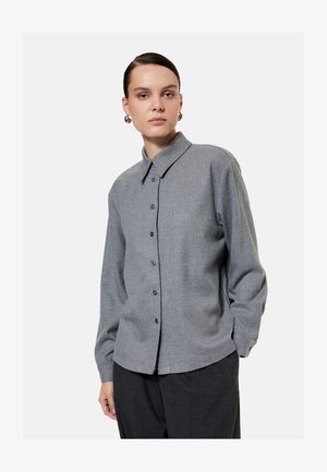 Mujer con el pelo peinado hacia atrás, lleva pendientes de aro plateados y una camisa gris de manga larga abotonada con pantalones gris oscuro sobre un fondo liso.