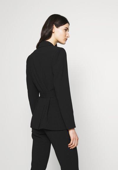 Blazer nero su misura con un design strutturato, colletto a punta, maniche lunghe e vita cinturata. Presenta una schiena plissettata per un dettaglio aggiuntivo.