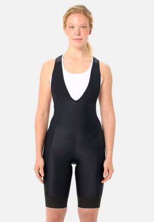 Donna con capelli chiari e ricci, che indossa pantaloncini da ciclismo neri con bretelle e una canotta bianca senza maniche, su uno sfondo bianco uniforme.