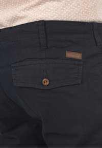 Pantalon en coton noir avec une seule poche arrière, fermeture par bouton marron et étiquette en tissu marron avec l'inscription "INDICODE." Texture douce et design décontracté.