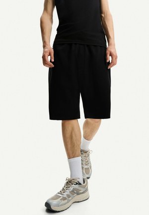 Man met zwarte korte broek, zwart mouwloos shirt, witte sportsokken en beige-grijze sneakers, staand op een witte achtergrond.