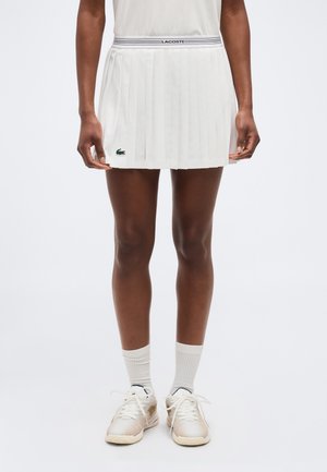 Osoba oblečená v bielej plisovanej teniskovej sukni Lacoste, bielych ponožkách a bielych teniskách stojaca pred jednoliatym pozadím.