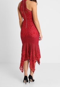 Frau, die ein ärmelloses rotes Spitzenkleid mit floralem Muster, einem asymmetrischen Saum und einem sichtbaren Reißverschluss im Rücken trägt, kombiniert mit schwarzen High Heels.