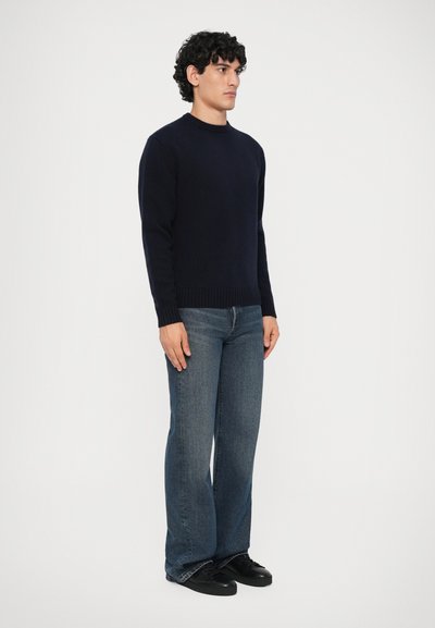 Giovane con capelli ricci che indossa un maglione blu navy, jeans blu scoloriti e scarpe da ginnastica nere, in piedi davanti a uno sfondo neutro.