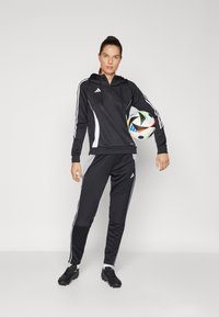 Black athletic hoodie with white accents and zip, paired with matching black pants and soccer ball. Model wearing black cleats.