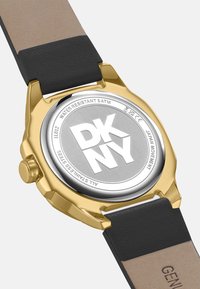 Goudkleurige roestvrijstalen watch achterkant met gegraveerd "DKNY." Zwarte leren band met tan accenten, met een kroon voor het instellen van de tijd.