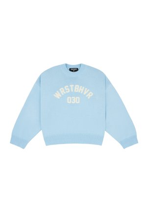 Femme portant un pull bleu clair oversize avec « WRSTBHVR 030 », un pantalon beige à jambes larges, des chaussures noires pointues, des lunettes de soleil, et un sac à main bleu clair.