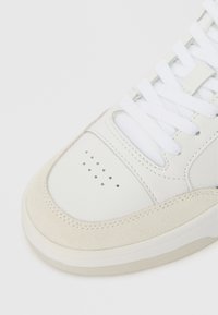 Λευκό sneaker σε κοντινό πλάνο που δείχνει την διάτρητη περιοχή του δαχτύλου, την σουέτ περιφέρεια και τα λευκά κορδόνια σε λευκό φόντο.