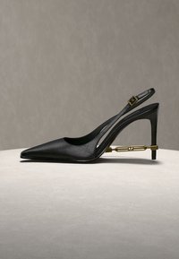 Black leather slingback high heel shoe with pointed toe and a gold metallic decorative support near the heel with "D2" engraving.