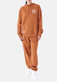 Conjunto de sudadera marrón y pantalones de chándal a juego en tejido suave. Presenta un bolsillo frontal, detalle de logo y puños elásticos. Se lleva con zapatillas blancas.