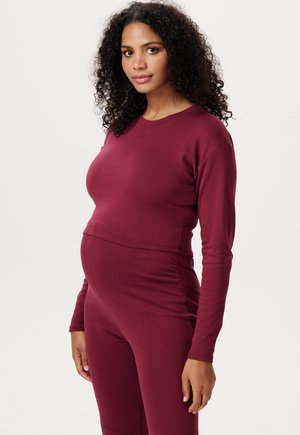 Zwangere vrouw met krullend haar, gekleed in een aansluitende bordeauxrode top met lange mouwen en bijpassende broek, staand tegen een effen lichte achtergrond.