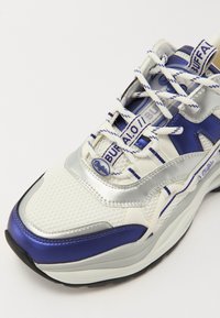 Hvite og sølvfargede joggesko med blå detaljer, laget av mesh-materiale, hevede såler, fremtredende snøringer og merkevarelogo på tungen og sidene.