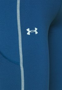Niebieskie legginsy sportowe o gładkiej teksturze, z białym logo Under Armour i kontrastowymi białymi szwami wzdłuż szwów.