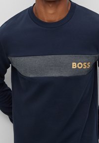 Marinblå långärmad t-shirt med en horisontell grå panel. Har en guldfärgad "BOSS"-logotyp på bröstet. Tillverkad av mjukt tyg med en enkel design.