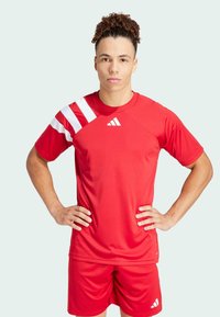 Rotes Sporttrikot und Shorts mit einem weißen diagonalen Streifen auf der Schulter, mit dem Adidas-Logo, das prominent auf der Brust abgebildet ist.