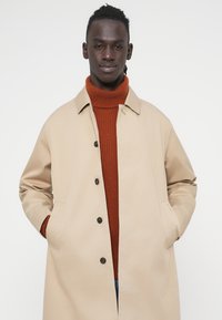 Μπεζ εξωτερικό παλτό με γιακά και κουμπιά μπροστά, φορεμένο πάνω από ένα πορτοκαλί ριγωτό ζιβάγκο. Διαθέτει πλαϊνές τσέπες και άνετη γραμμή.