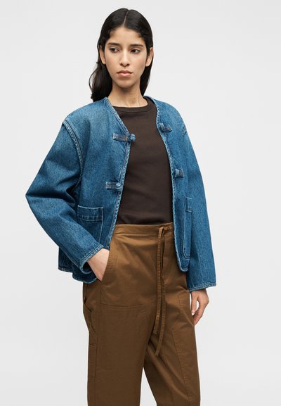 Τζιν μπουφάν με μπλε σώμα, ανοιχτό γιακά και κλεισίματα με toggle. Φορεμένο πάνω από καφέ πουκάμισο και συνδυασμένο με καφέ στενά παντελόνια.