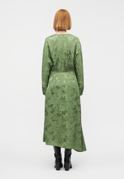 Μακρύ, πράσινο φόρεμα από απαλό ύφασμα με ανάγλυφο μοτίβο. Μακριά μανίκια και ραφή στην πλάτη, σε συνδυασμό με μαύρες μπότες μέχρι το γόνατο.
