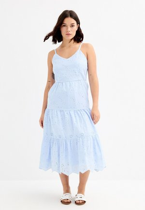 Vestido de algodón azul claro con un diseño en capas, patrones de ojal, tirantes finos y un dobladillo con forma de ondulación. Llevado con sandalias blancas de tiras.