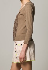 Brązowy dziergany kardigan z teksturowanymi falbankami z przodu, zestawiony z kremową spódnicą ozdobioną kolorowym haftem i neonowymi akcentami.