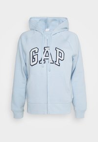 Light blue zip-up hoodie with large "GAP" logo, front pockets, and a drawstring hood. Made from soft, textured fabric.