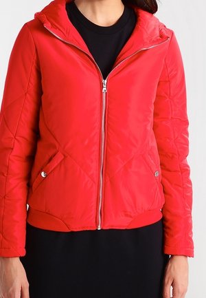 Vrouw draagt een rode doorgestikte jas met ritssluiting en zijzakken over een zwart shirt, afgebeeld van schouders tot heupen.