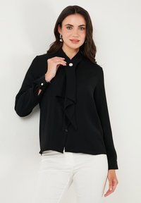 Black blouse made of smooth fabric featuring a bow tie neckline, long sleeves with button cuffs, and a slim fit design.