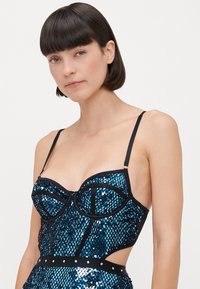 Top en sequins bleus avec bord noir, présentant un design de bustier structuré et des bretelles réglables, rehaussé d'une texture en maille et de clous argentés.
