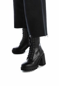 Botas de cuero negras hasta el tobillo con un tacón alto y grueso, cordones delanteros y suela texturizada, con ojales de metal y costuras decorativas.