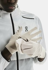 Gants beiges légers fabriqués en matériau lisse et élastique. Présentent un ajustement snug et des bouts de doigts arrondis, avec un détail de logo sur le poignet.
