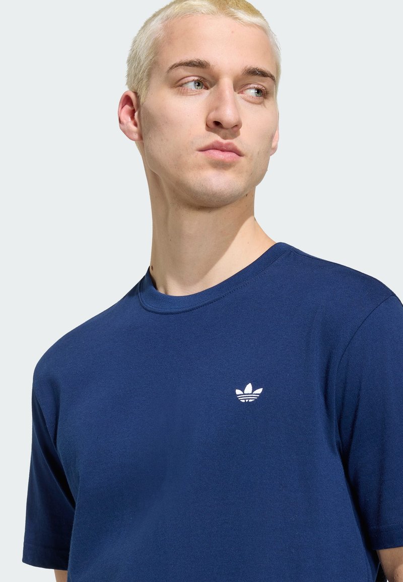 Jeune personne aux cheveux blonds courts portant un t-shirt Adidas bleu marine, regardant sur le côté devant un fond clair uni.