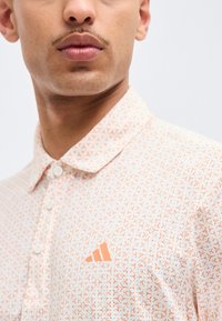 Κοντινό πλάνο ατόμου που φοράει λευκό πόλο πουκάμισο με πορτοκαλί γεωμετρικό σχέδιο και πορτοκαλί λογότυπο Adidas στο στήθος.