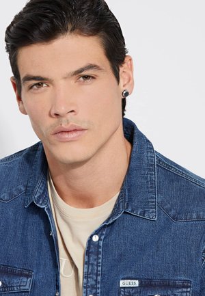 Chaqueta de mezclilla azul con un acabado texturizado, dos bolsillos en el pecho, acentos de botón blanco y una camiseta beige clara debajo.