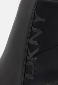 Gros plan d'un panneau de chaussure en cuir et tissu noir avec le logo noir brillant « DKNY » embossé verticalement.