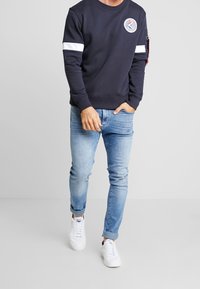 Sweat-shirt navy avec des rayures blanches sur les manches, écusson logo sur la poitrine, associé à un jean slim bleu clair et des baskets blanches.