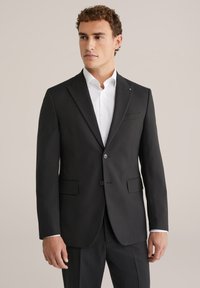 Melnā piegriezuma uzvalka žakete ar šauru piegriezumu, vienu pogu, izgrieztām apkaklēm un divām priekšējām kabatām. Zem tās balta krekliņa.