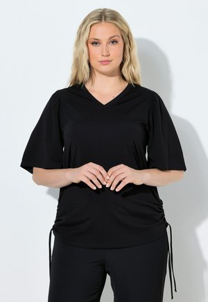 Kvinde med langt blondt hår iført en sort V-hals top med korte puffede ærmer og snor i siderne, stående mod en hvid baggrund.