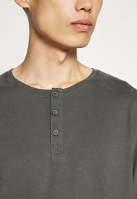 Inimese lähivaade, kes kannab tumehallit henley-kujulist särki, millel on kolm nööpi, näidates kaela ja osaliselt külje peale pööratud nägu.