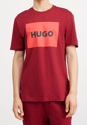 Czerwony bawełniany T-shirt z prostokątnym nadrukiem w kolorze koralowym i napisem „HUGO” pogrubioną czarną czcionką na przodzie.