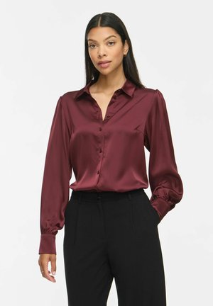 Femme aux longs cheveux foncés portant un chemisier en satin bordeaux boutonné et un pantalon noir, debout avec une main dans la poche.