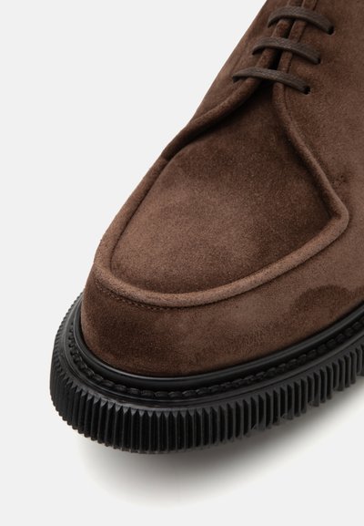 Brown suede shoe with a rounded toe and raised stitching detail. Features a black textured rubber sole with ridges for grip.