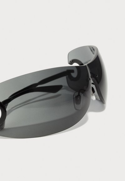 Saulesbrilles ar tumši pelēkiem, ietverošiem stikliem un izliekto rāmi. Aprīkotas ar elegantiem melniem uzgaļiem un apaļiem akcentiem pie templiem.