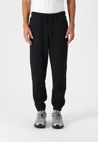 Zwarte trainingsbroek van zacht stof, met een elastische tailleband en trekkoord, taps toelopende enkels en zijzakken. Draagt met grijze sportschoenen.